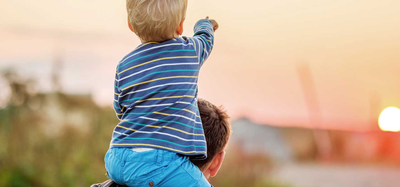 Young boy pointing at the sunset while sitting on his father's shoulders.
