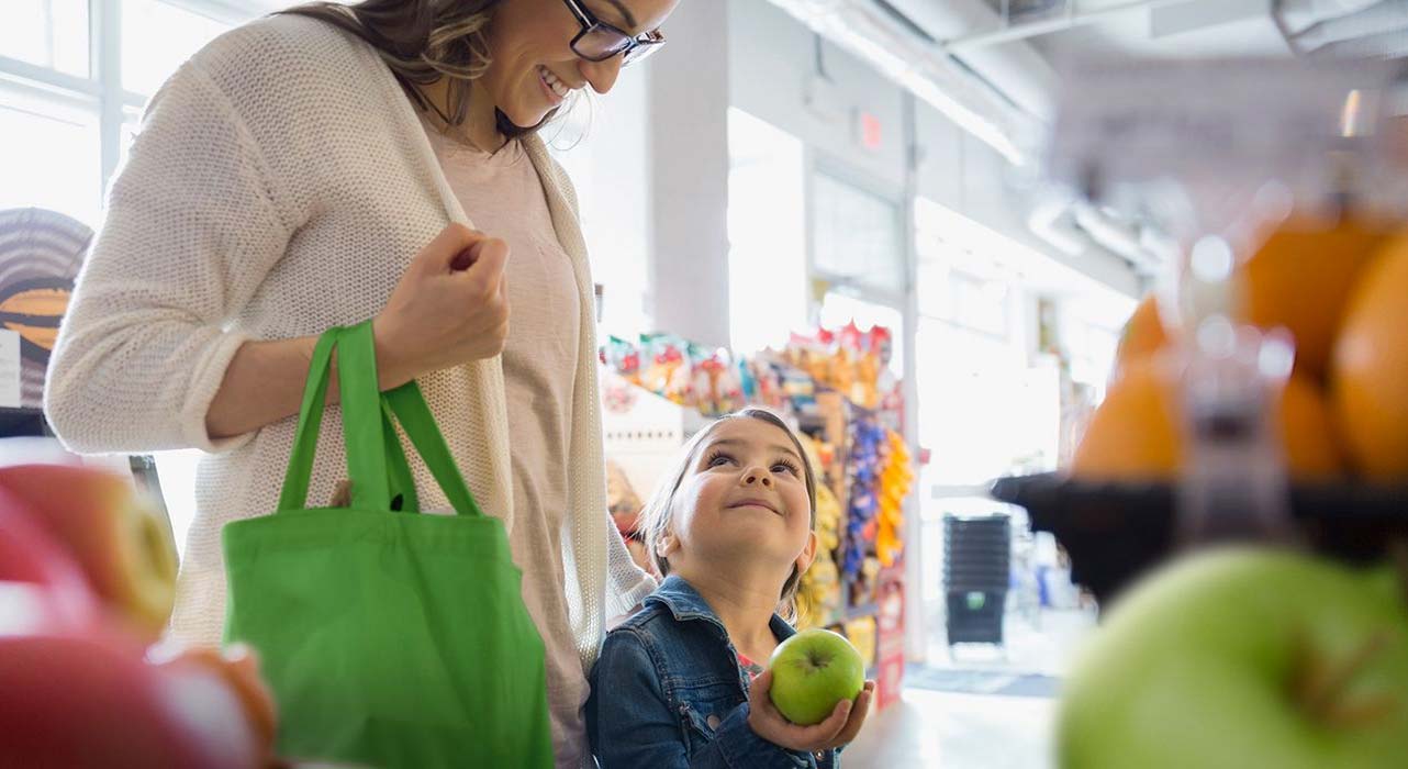 A little girl holding an apple, looking up at her mother in a grocery store.