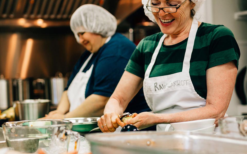 Deux femmes avec un tablier qui pèlent des carottes dans une cuisine.