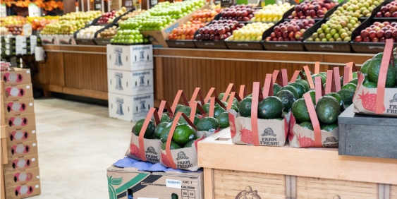 A grocery store display featuring various fruits, with avocados in the foreground and neatly arranged apples and other fruits in the background.